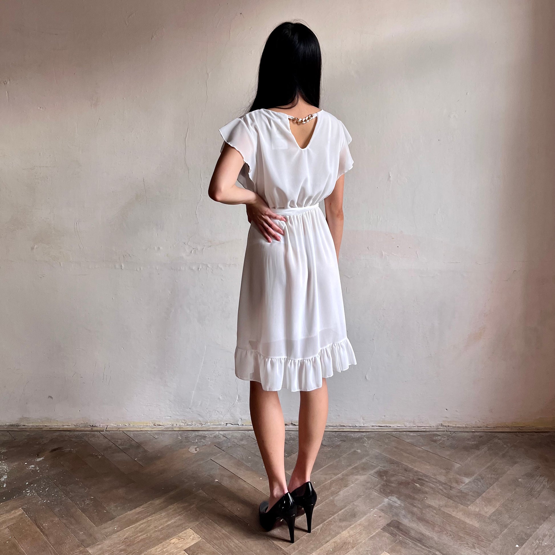 Modelka asijského původu pózující zezadu oblečená ve krátkých bílých společenských šatech s volány