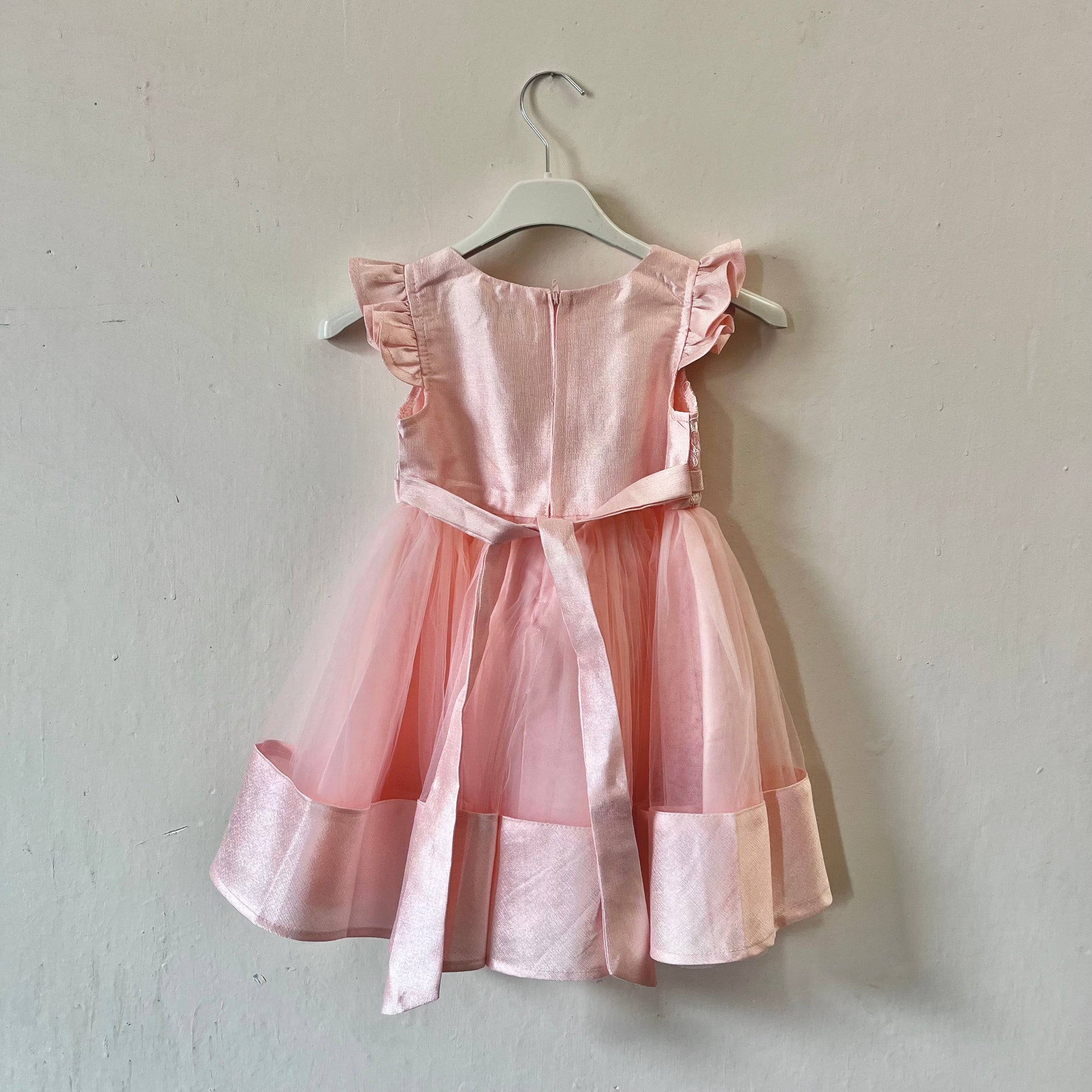 Dívčí růžové krajkované šaty s mašlí visící na věšáku přední stranou ke zdi