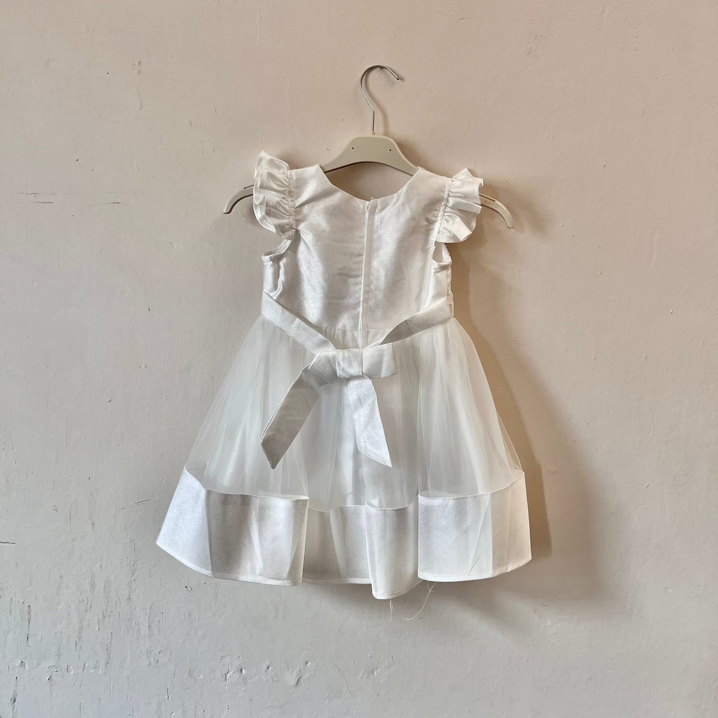 Dívčí bílé krajkované šaty s mašlí visící na věšáku přední stranou ke zdi